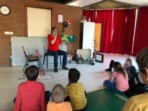 Plusieurs enfants sont assis en cercle et écoutent une dame habillée en rouge raconter une histoire et faire le son d'un oiseau.