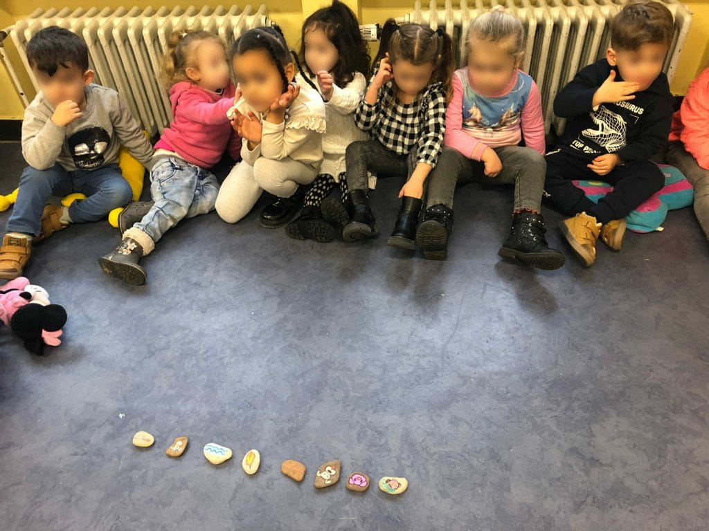 Des enfants de maternelle sont assis par terre en cercle et regardent des petits cailloux sur lesquels se trouvent des dessins faits à la peinture.
