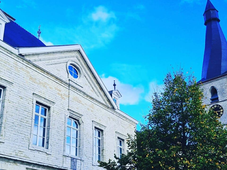 Photo de l'Hôtel des Libertés et de la Chapelle Notre-DAme du Marché, deux bâtiments historiques situés à Jodoigne. Le ciel est bleu vibrant et un arbre se trouve au premier plan.