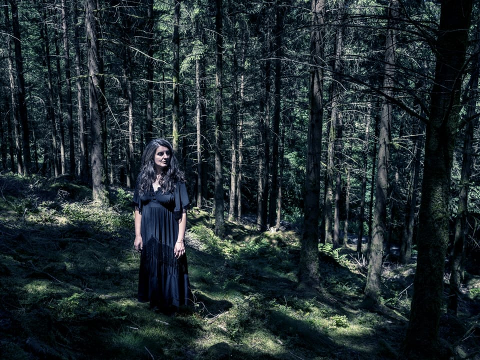 La conteuse Catherine Pierloz se trouve dans une forêt assez sombre où les rayons du soleil réussissent à percer par endroit.